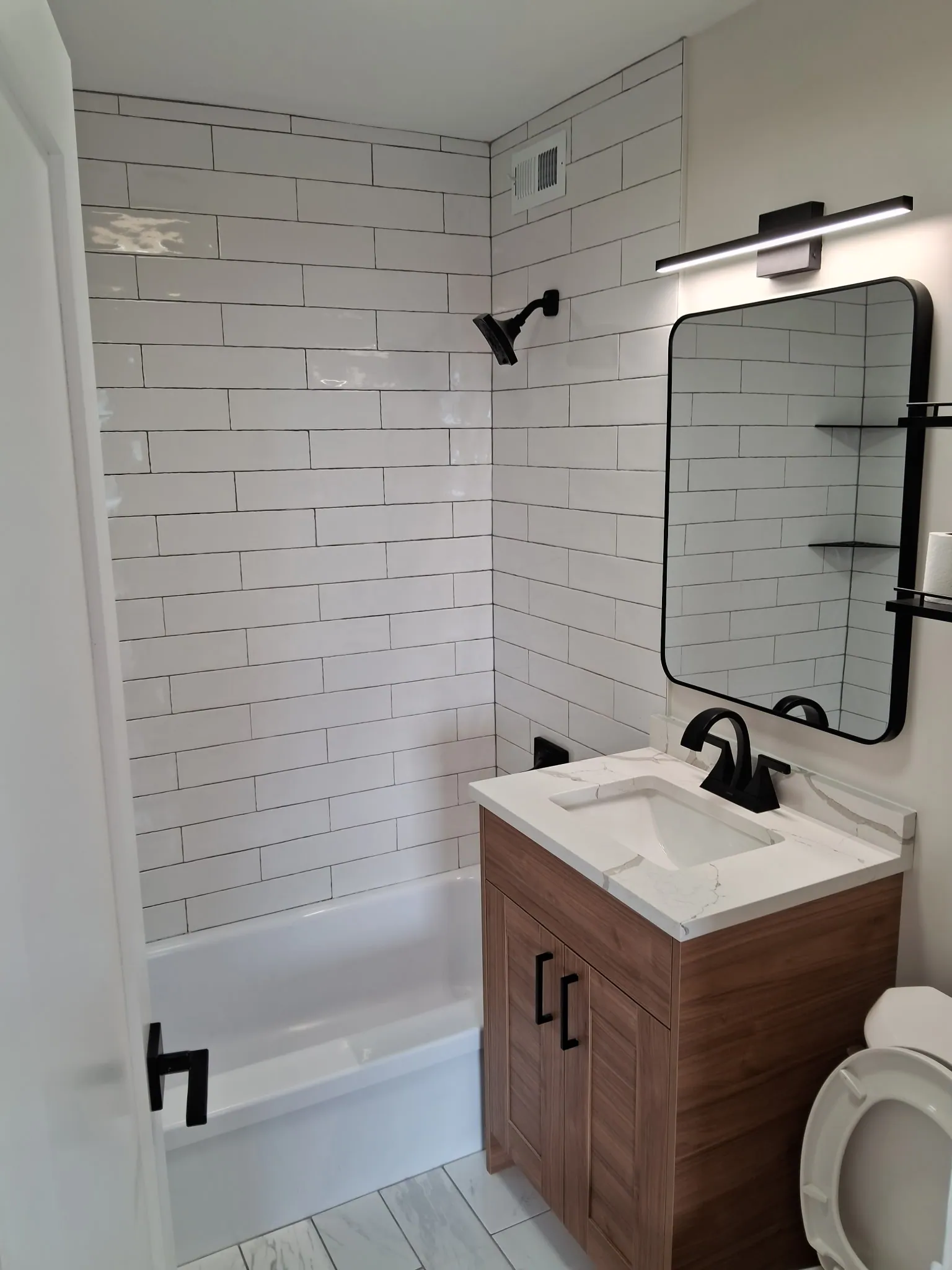 Remodeled condo bathroom with white subway tile tub-shower, wood-tone vanity, black fixtures and a black-framed mirror